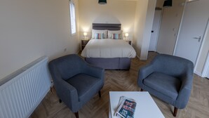 A bedroom with a bed, two armchairs, a bedside table with a magazine, a radiator, and a window with blinds. - Wick Bay Apartments (Wick)