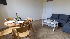 A dining area with a round wooden table, four chairs, and a bottle of wine. There is a grey sofa and a white coffee table in the living area. - Wick Bay Apartments (Wick)