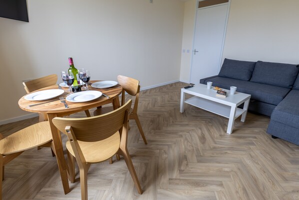 A dining area with a round wooden table, four chairs, and a bottle of wine. There is a grey sofa and a white coffee table in the living area. - Wick Bay Apartments (Wick)
