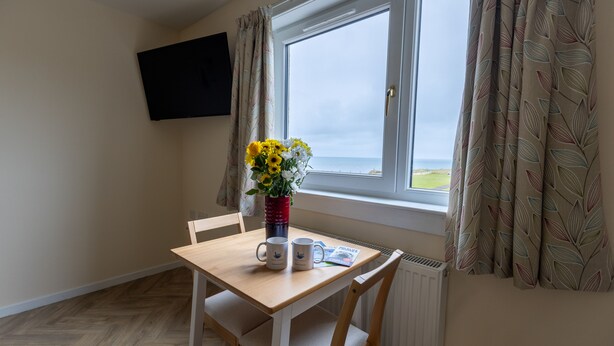 A room with a wooden table, two chairs, a vase with flowers, two mugs, and a flat-screen TV mounted on the wall. - Wick Bay Apartments (Wick)