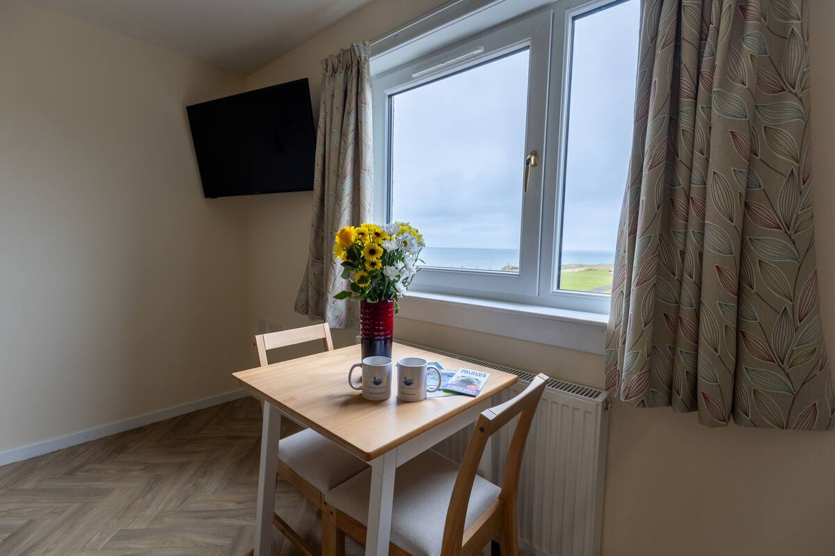 A room with a wooden table, two chairs, a vase with flowers, two mugs, and a flat-screen TV mounted on the wall.