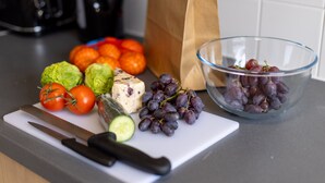 A kitchen counter with a cutting board, a knife, tomatoes, cucumbers, cheese, grapes, and a paper bag. - Wick Bay Apartments (Wick)