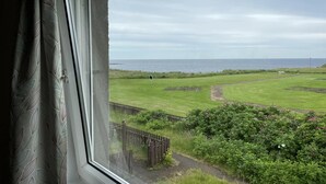 A view through a window of a grassy area with a wooden fence, a path, and a body of water in the distance. - Wick Bay Apartments (Wick)