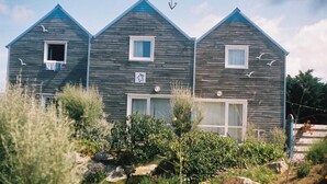 A row of three wooden houses with white trim and windows, surrounded by greenery and rocks. - Gîte Cozy à Deux pas de la mer (Le Conquet)