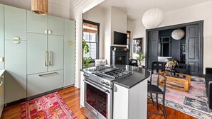 Una cocina moderna con una isla central, un comedor con sofá negro y mesa de madera, y una sala de estar con chimenea y paredes negras.