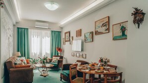 A cozy living room with a sofa, armchairs, a dining table set with plates and cups, and a coffee table. There are curtains, wall art, and a clock on the wall. - pnoe d'Athènes (Athina)