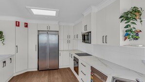 A modern kitchen with white cabinetry, a stainless steel refrigerator, a microwave, and a dishwasher. There are ceiling lights and a plant on the wall. - Merimbula Heights 1 - Merimbula (Merimbula)