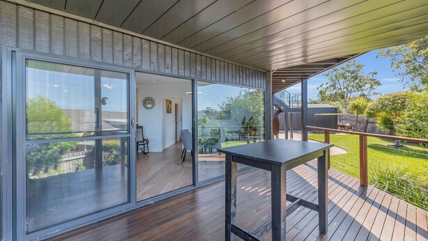 A modern outdoor deck with a wooden table and chairs, large sliding glass doors leading to a patio area, and a view of the surrounding greenery.