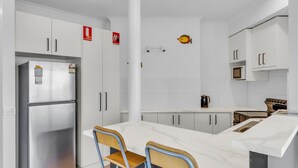 A modern kitchen with white cabinets, a stainless steel refrigerator, a microwave, and a stove. There are two wooden chairs at a white marble table.