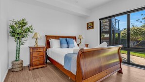 A bedroom with a wooden bed, two bedside tables with lamps, a large window with a view of the outdoors, and a potted plant.