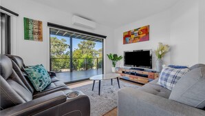 A modern living room with a large TV, comfortable seating, a coffee table, and a balcony with a view of trees.