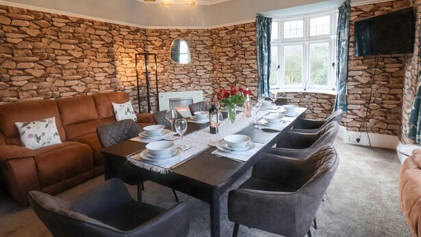 Un espace repas agr émenté d’un mur en pierre, d’une cheminée, d’une longue table dressée pour six personnes et d’un coin salon confortable.