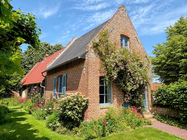 A brick house with a steeply pitched roof, a visible chimney, and a garden with various plants and flowers.