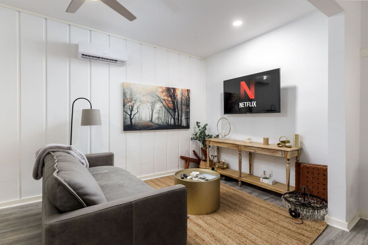 Una sala de estar moderna con un sofá gris, una mesa de centro redonda dorada, un televisor montado mostrando el logotipo de Netflix, un cuadro en la pared y un mueble consola de madera con objetos decorativos.