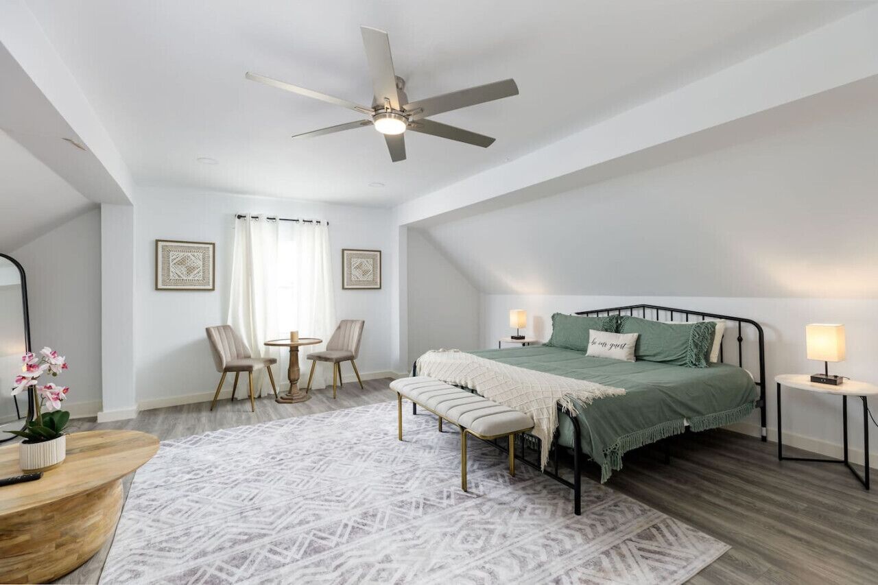 A bedroom with a bed, a ceiling fan, a bedside table with a lamp, a bench, a chair, a small table, and a window with curtains.