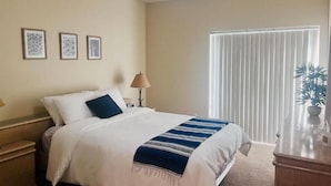 Habitación de hotel con cama, mesita de noche, lámpara, ventana con persianas y una planta.