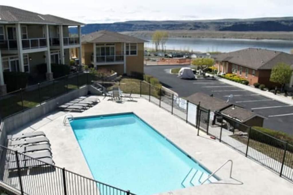 A swimming pool with lounge chairs, a parking area, and buildings in the background.