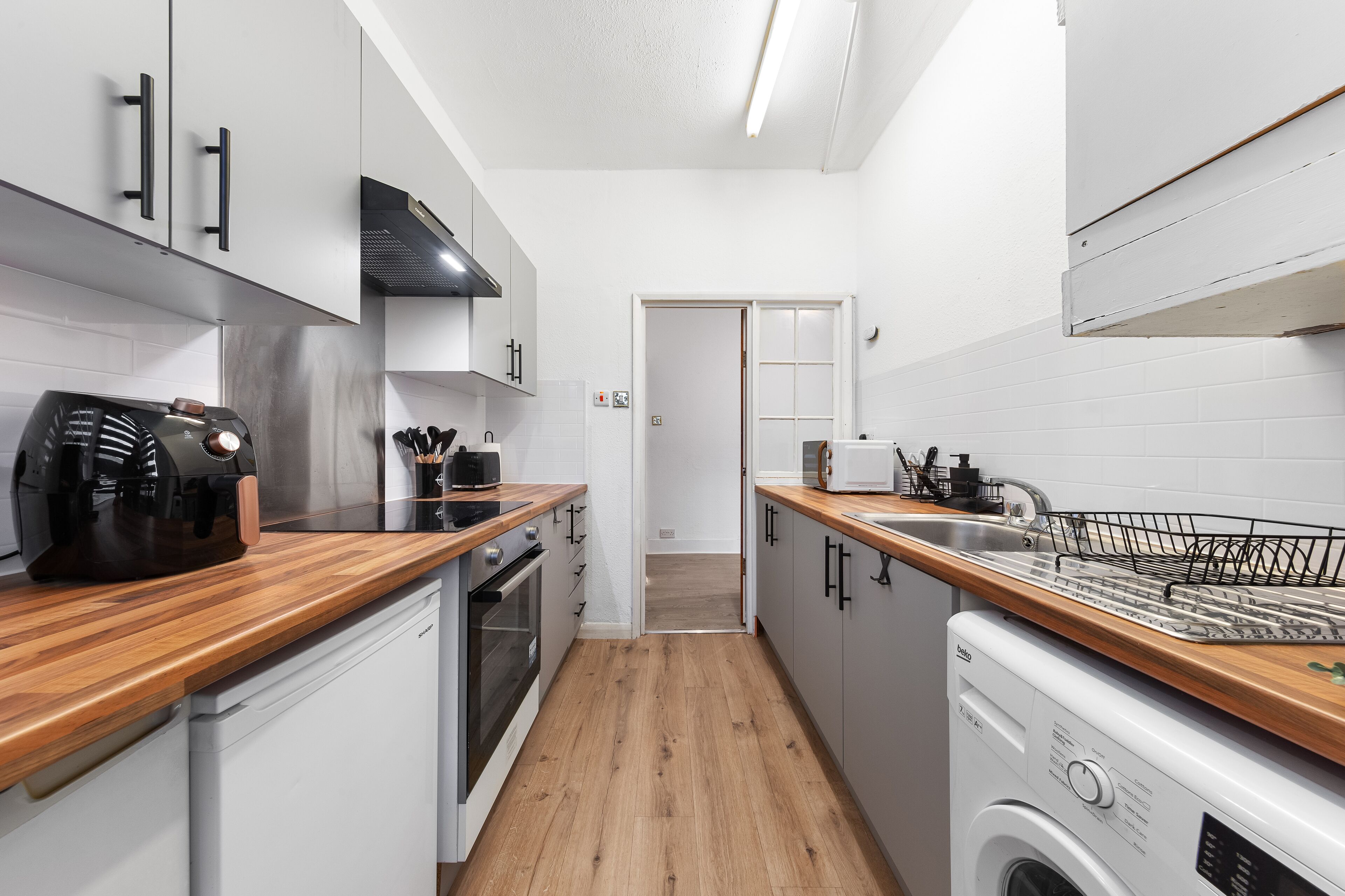 Une cuisine compacte avec des meubles blancs, un plan de travail en bois, un four encastré, un lave-vaisselle et un four à micro-ondes.
