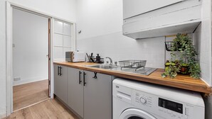 A compact kitchen with a washing machine, a sink, and a countertop with a plant. - Elegant 1 Bdr Flat in Goodmayes w Free Parking (Ilford)