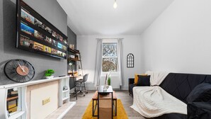 A modern living room with a flat-screen TV mounted on the wall, a black sofa, a white armchair, and a yellow rug. - Elegant 1 Bdr Flat in Goodmayes w Free Parking (Ilford)