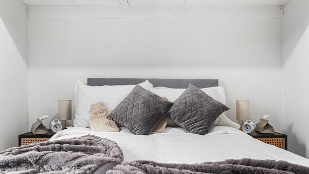 A neatly made bed with a gray quilted blanket, two pillows, and a bedside table with a lamp and a clock. - Elegant 1 Bdr Flat in Goodmayes w Free Parking (Ilford)