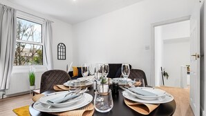 A dining area with a round table set for six, white plates, clear glasses, and a central vase. - Elegant 1 Bdr Flat in Goodmayes w Free Parking (Ilford)