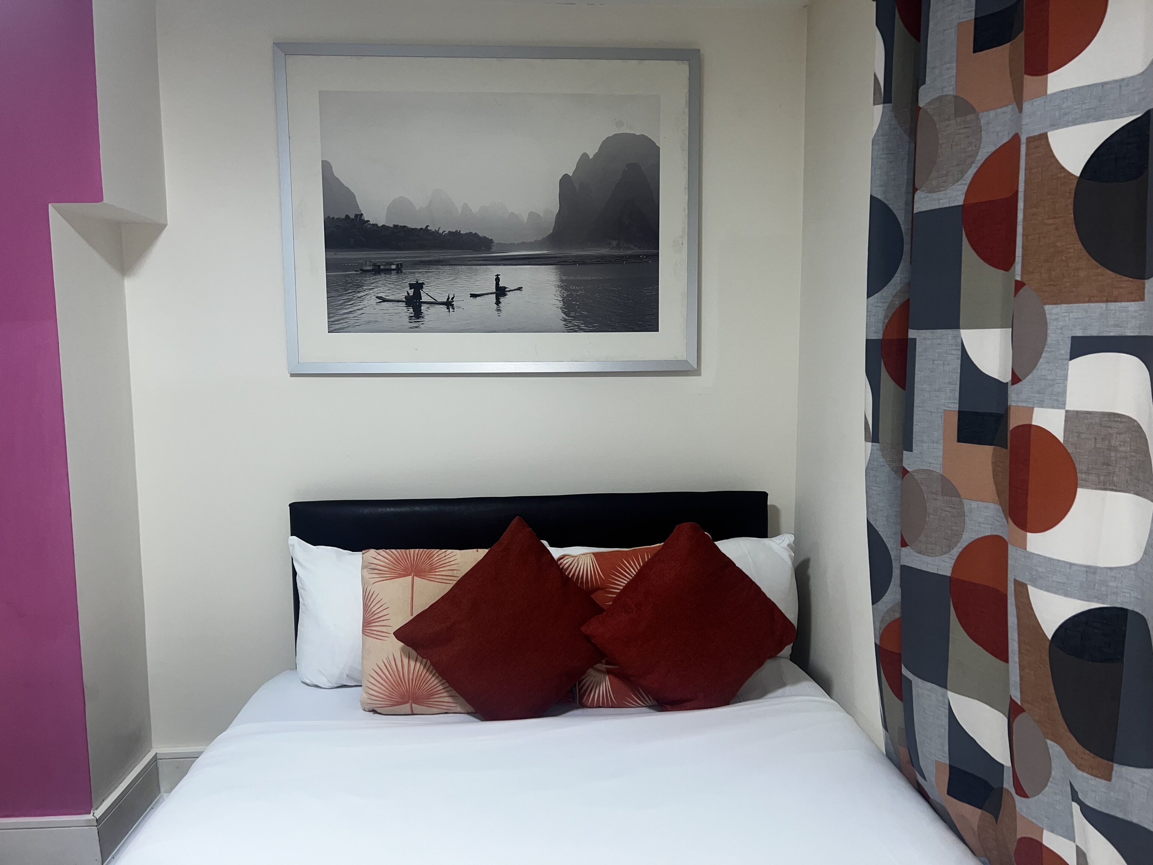 A neatly made bed with two decorative pillows, a framed picture of a landscape on the wall, and a patterned curtain.