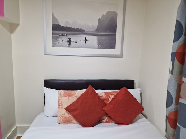 A bed with white bedding and two red pillows, a framed picture of a river scene above it, and a curtain with a circular pattern to the right.