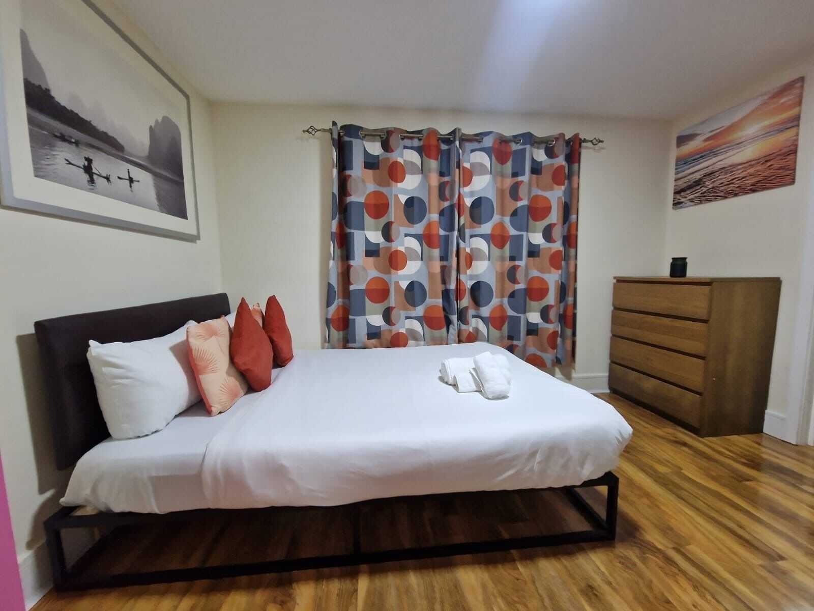 A neatly made bed with pillows, a wooden dresser, and a painting on the wall.
