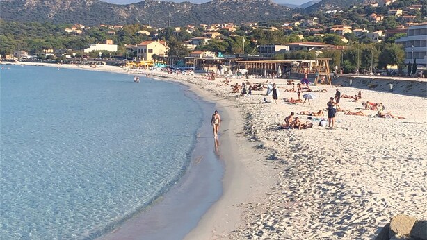 Ein Strand mit klarem Wasser, sandigem Ufer und Gebäuden im Hintergrund.