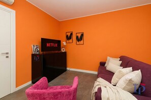 A room with orange walls, a purple sofa, a pink chair, and a TV on a stand.