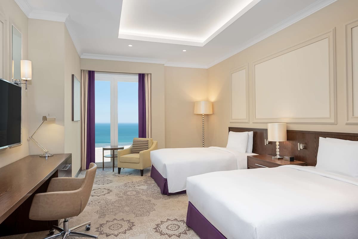 Quarto de hotel com duas camas, uma escrivaninha, uma cadeira, uma televisão e vista para o oceano.
