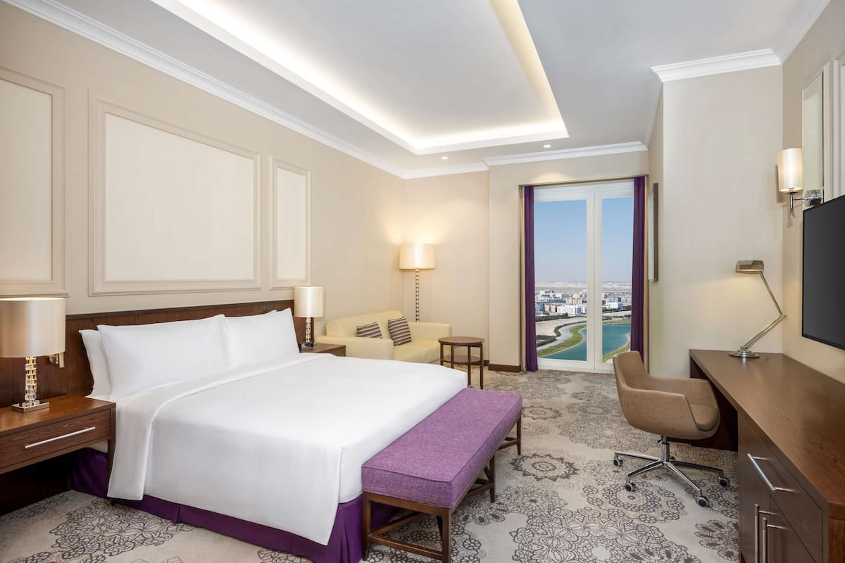 Quarto de hotel com uma cama grande, uma escrivaninha, uma cadeira, uma mesinha e vista para a cidade através de uma janela ampla.