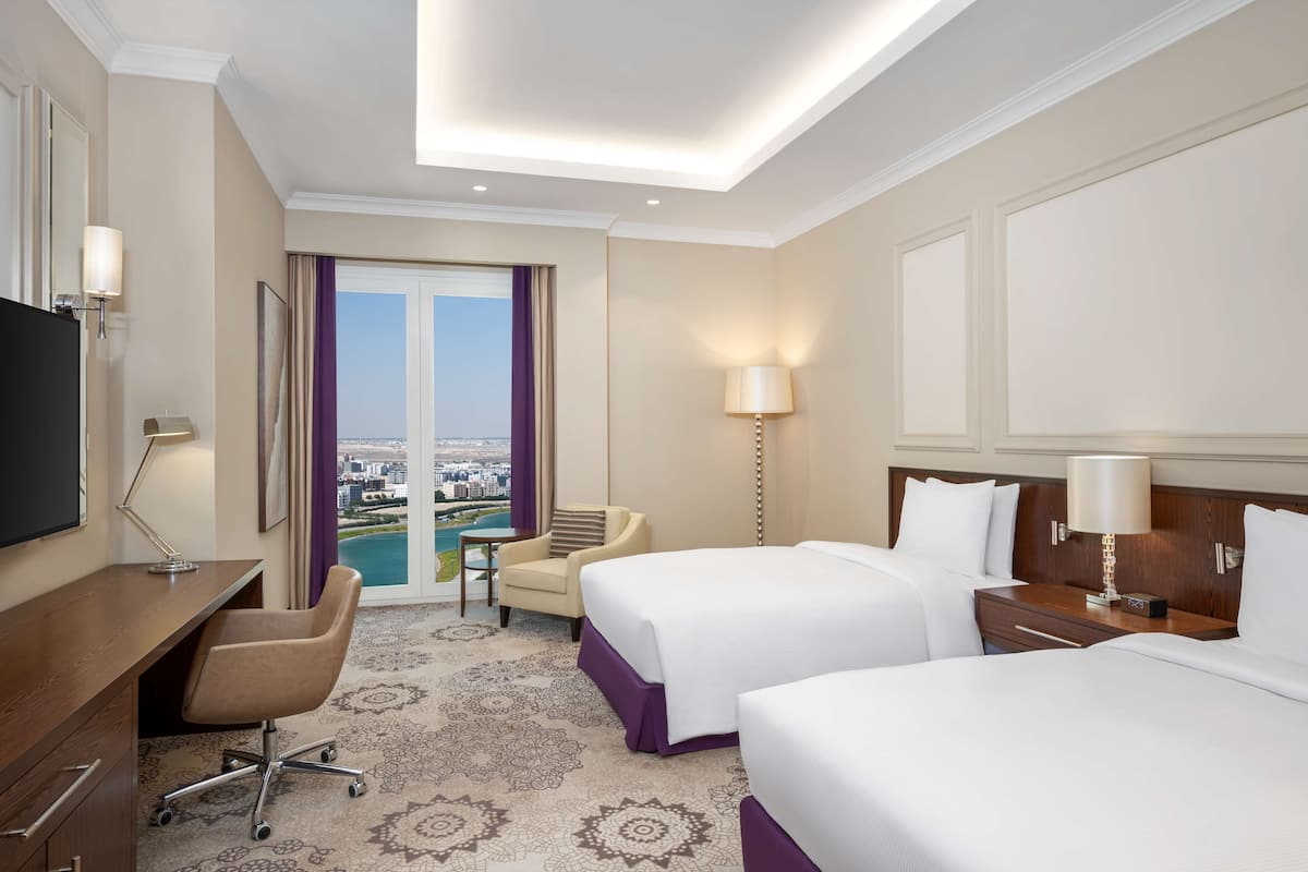 Quarto de hotel com duas camas, uma escrivaninha, uma cadeira, televisão e vista para a cidade.