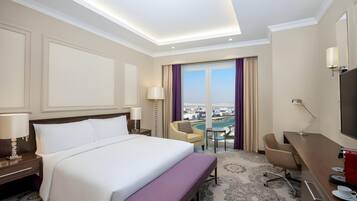 A hotel room with a large bed, a desk, a chair, and a view of a cityscape through the window.