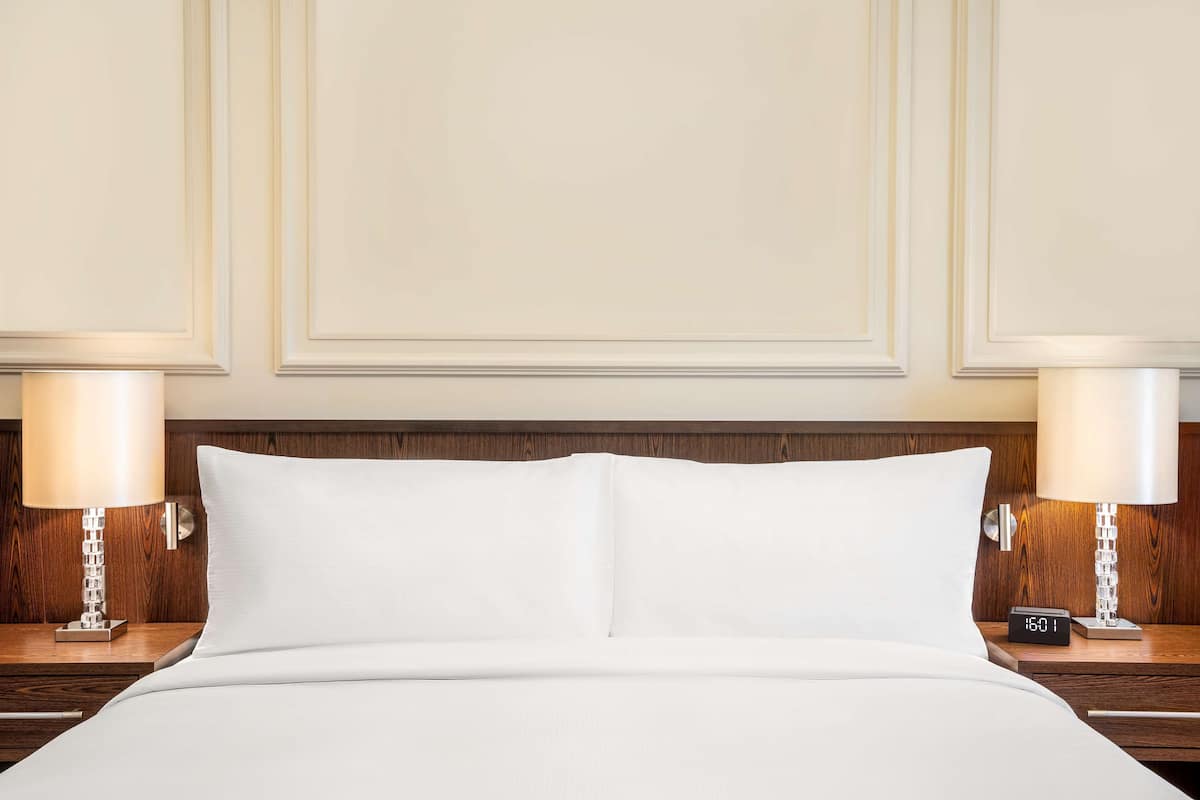 Cama bem arrumada com lençóis brancos, duas luminárias laterais e um relógio digital na mesa de cabeceira.
