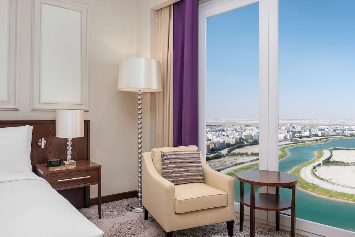 Quarto de hotel com cama, mesa de cabeceira com abajur, poltrona com almofada, mesinha e uma grande janela com vista para a cidade e para o rio.