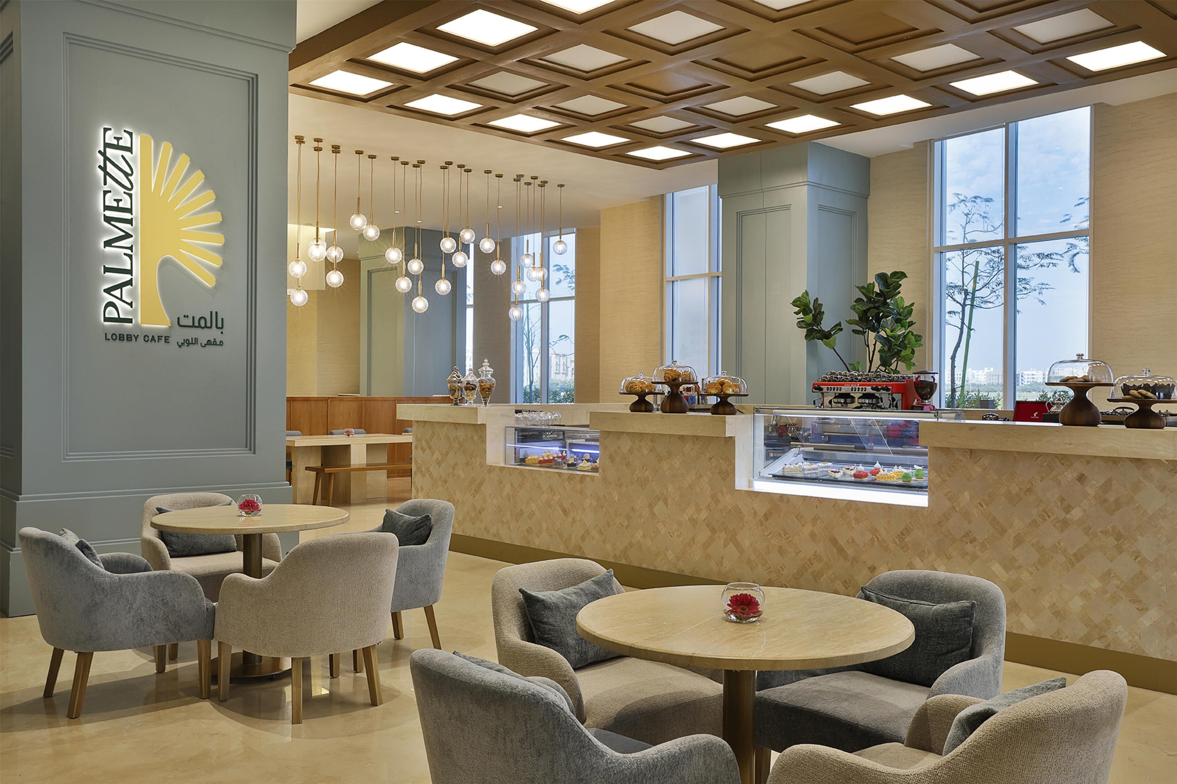 A lobby cafe with round tables and grey upholstered chairs, a decorative ceiling, and a view through large windows.