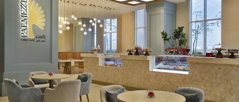 A lobby cafe with round tables and grey upholstered chairs, a decorative ceiling, and a view through large windows.