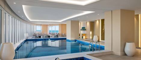 Un moderno vestíbulo de hotel con una amplia piscina cubierta, un jacuzzi independiente y un área de descanso con vista al mar.