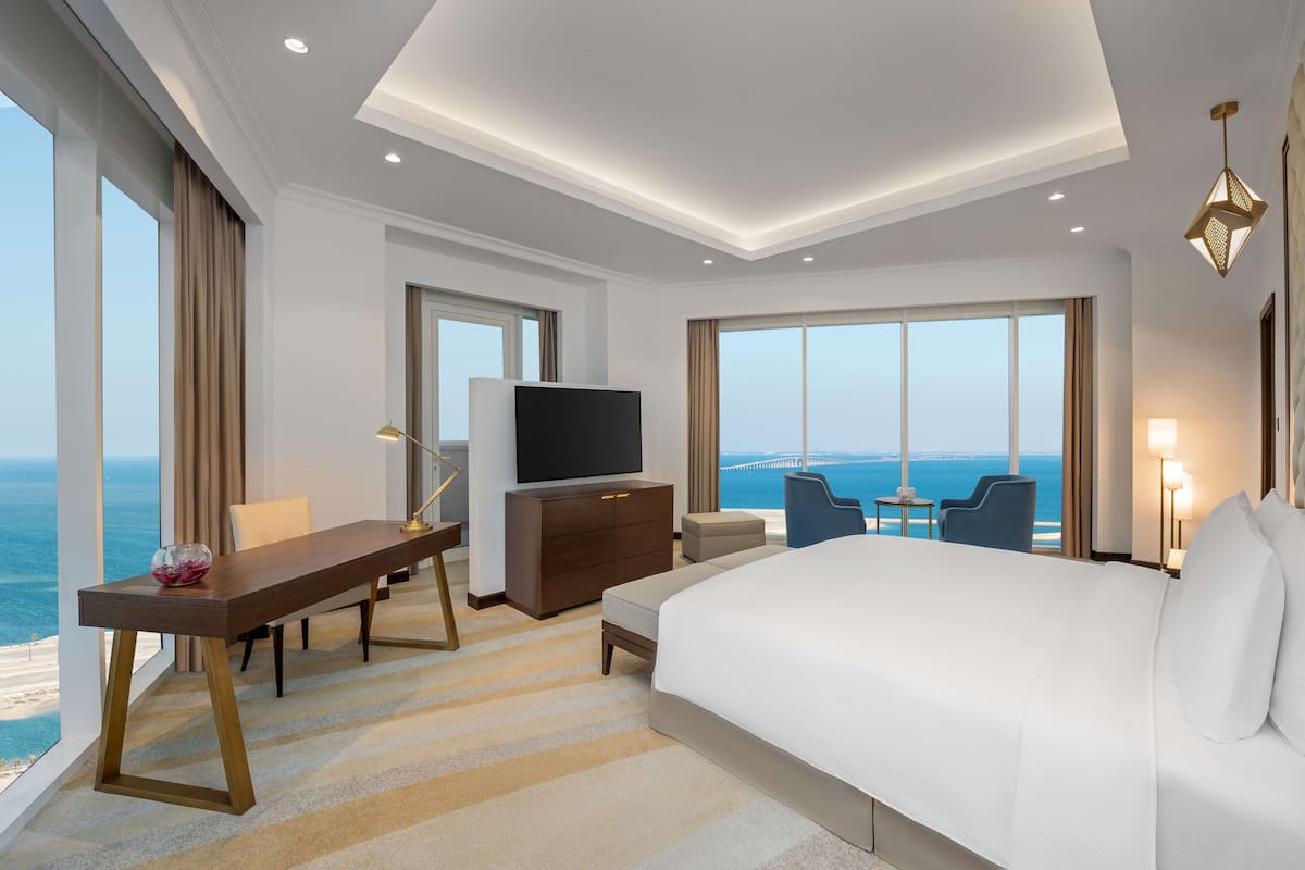 Quarto de hotel com uma cama grande, uma escrivaninha com televisão, vista para o oceano e varanda com mesa e cadeiras.