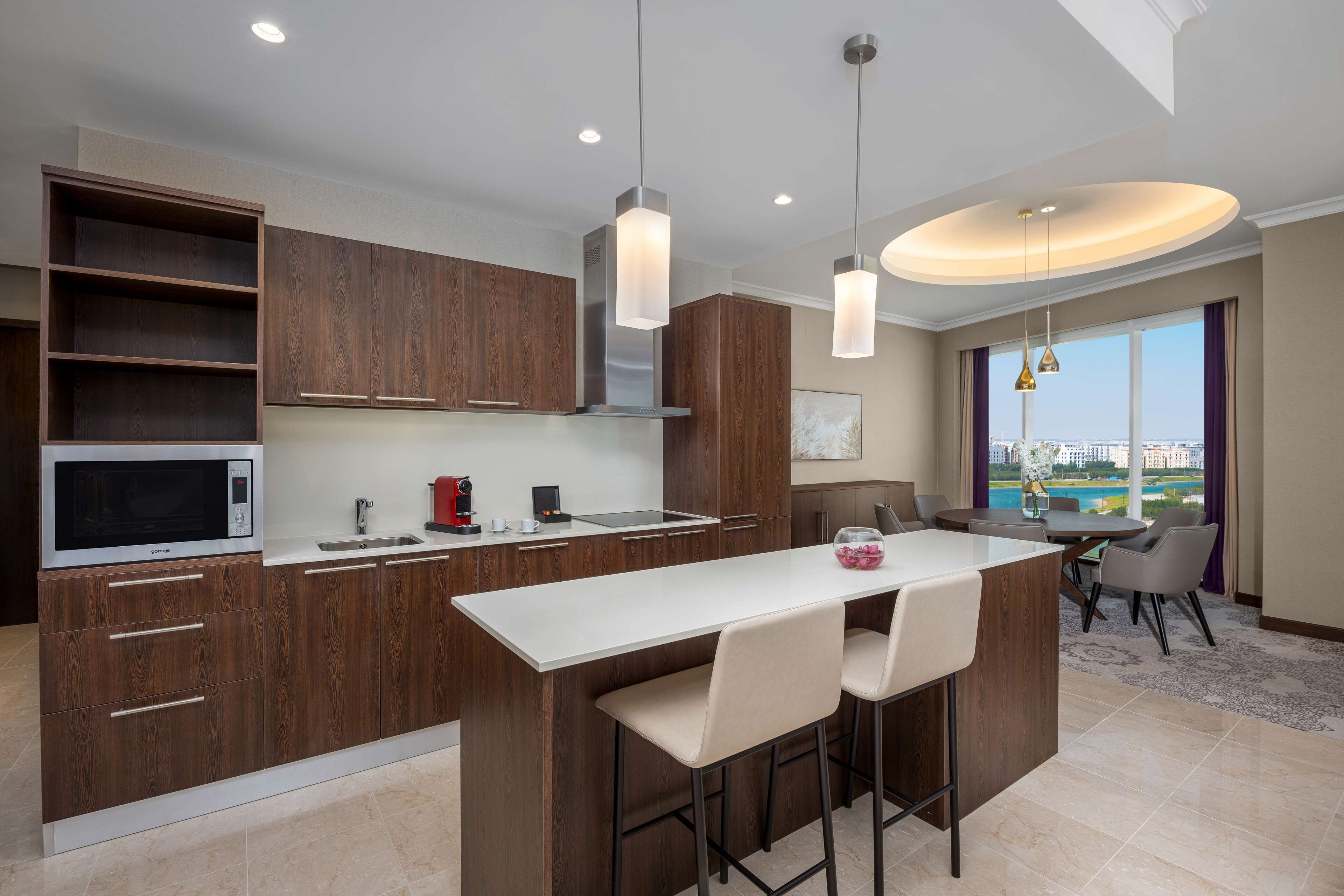 Una cocina moderna con una isla central, microondas integrado y campana extractora de acero inoxidable. Hay lámparas colgantes sobre la isla y un ventanal con vistas.