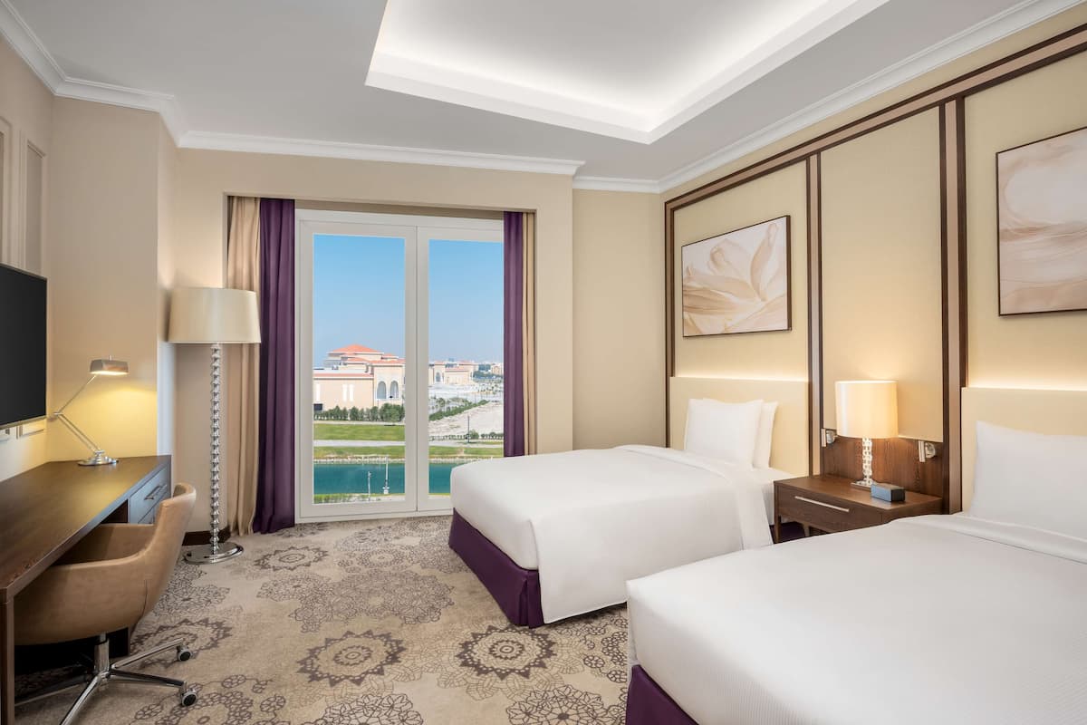 Quarto de hotel com duas camas, uma escrivaninha, uma cadeira, televisão e vista para um edifício e uma piscina.
