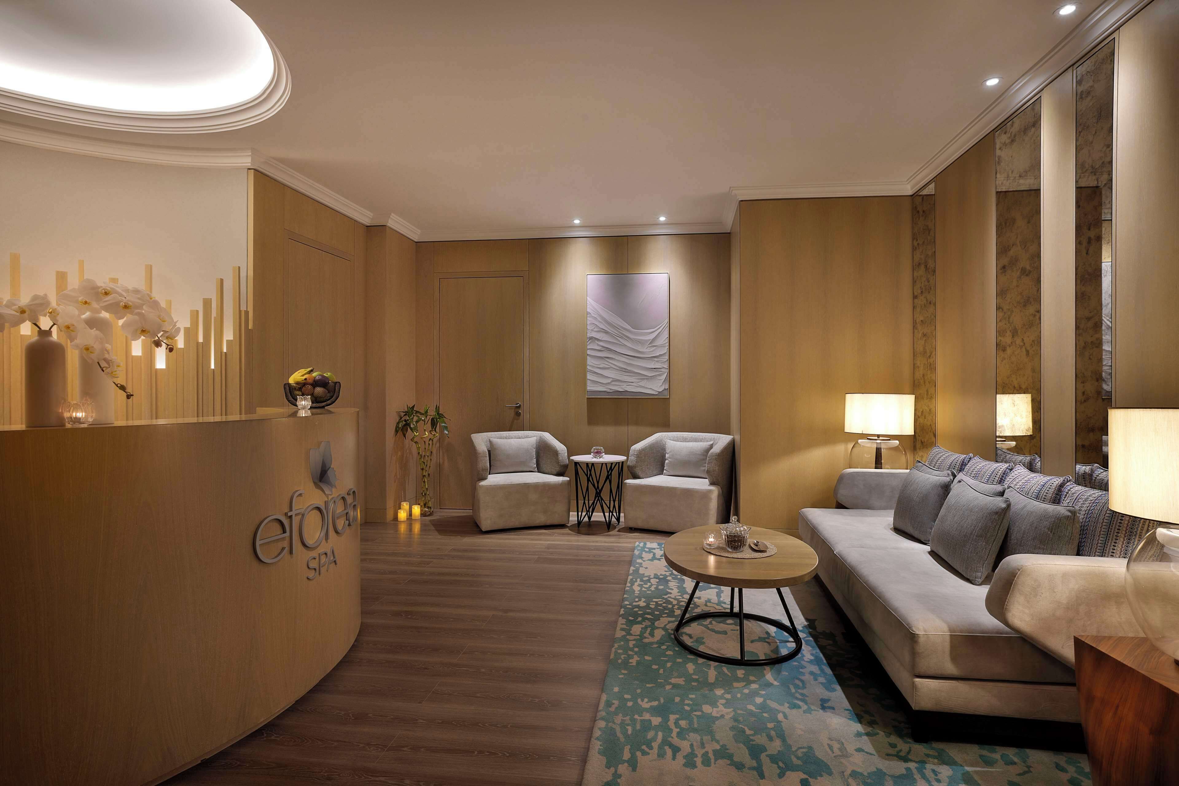 Een moderne spa-ontvangstzone met een gebogen receptiebalie, twee banken, een klein rond tafeltje en een grote spiegel.