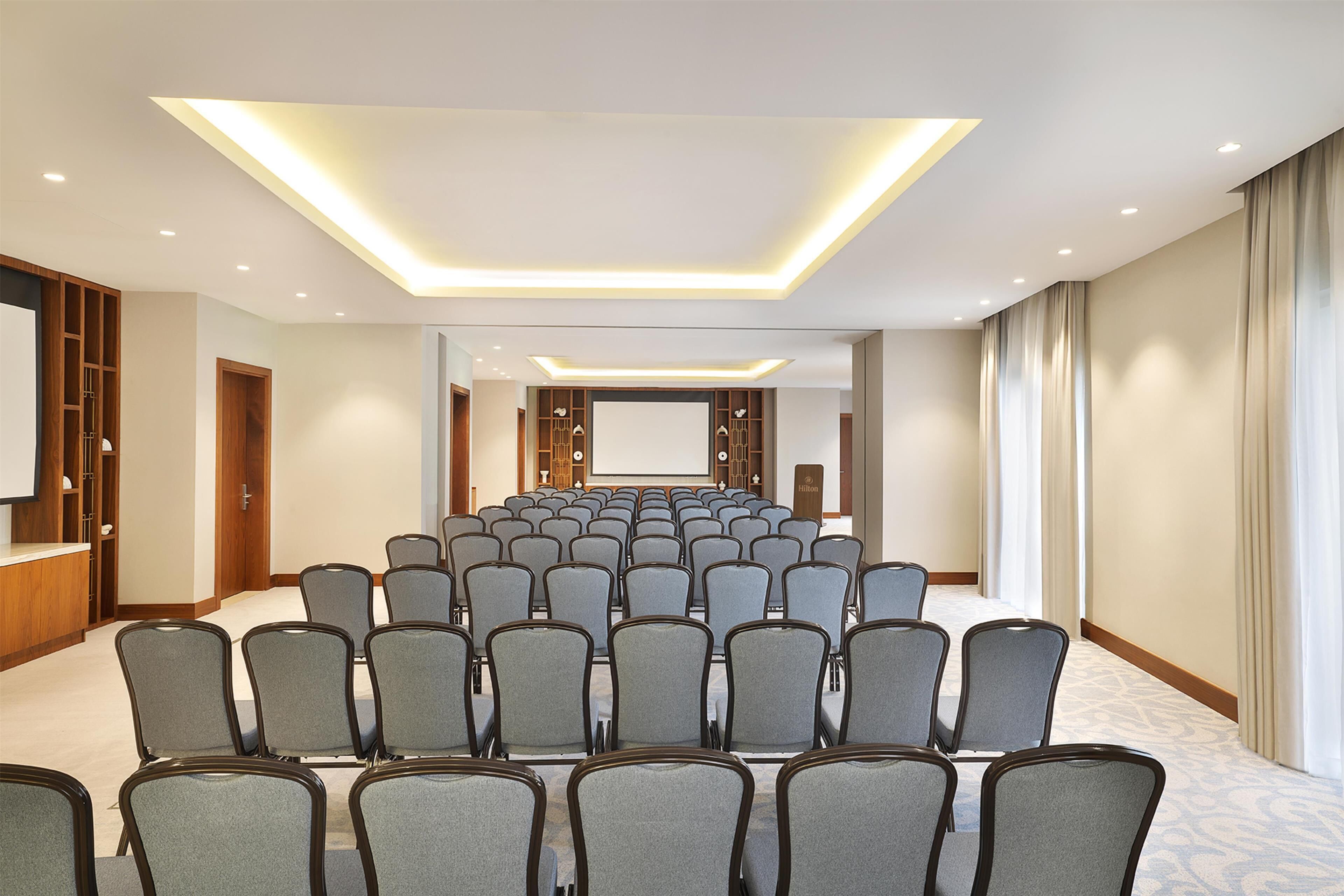 Un salón de conferencias con filas de sillas, una pantalla de proyector y una pantalla grande.