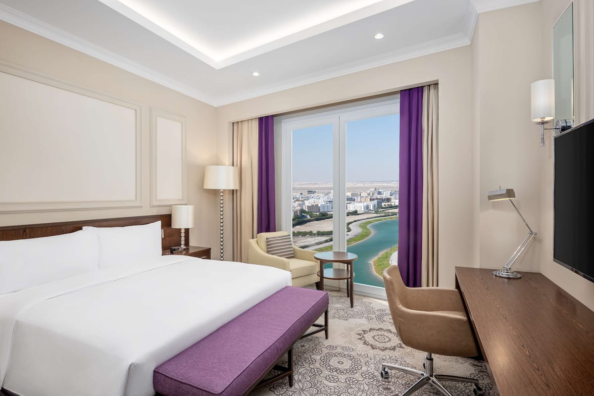 Quarto de hotel com uma cama grande, uma escrivaninha, uma cadeira, uma mesinha e vista para a cidade através de amplas janelas.