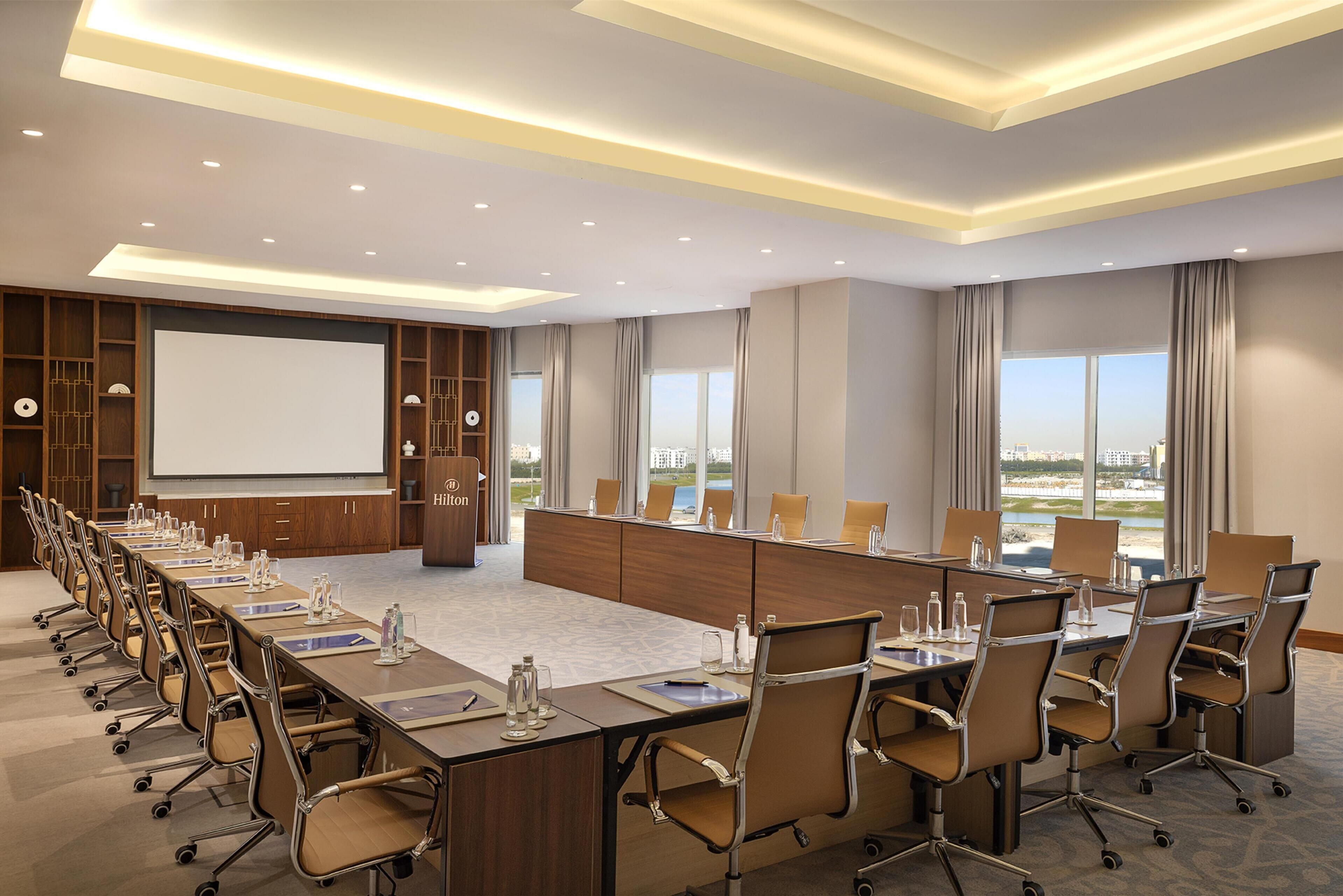 A conference room with a long table, chairs, a podium, and a large screen.