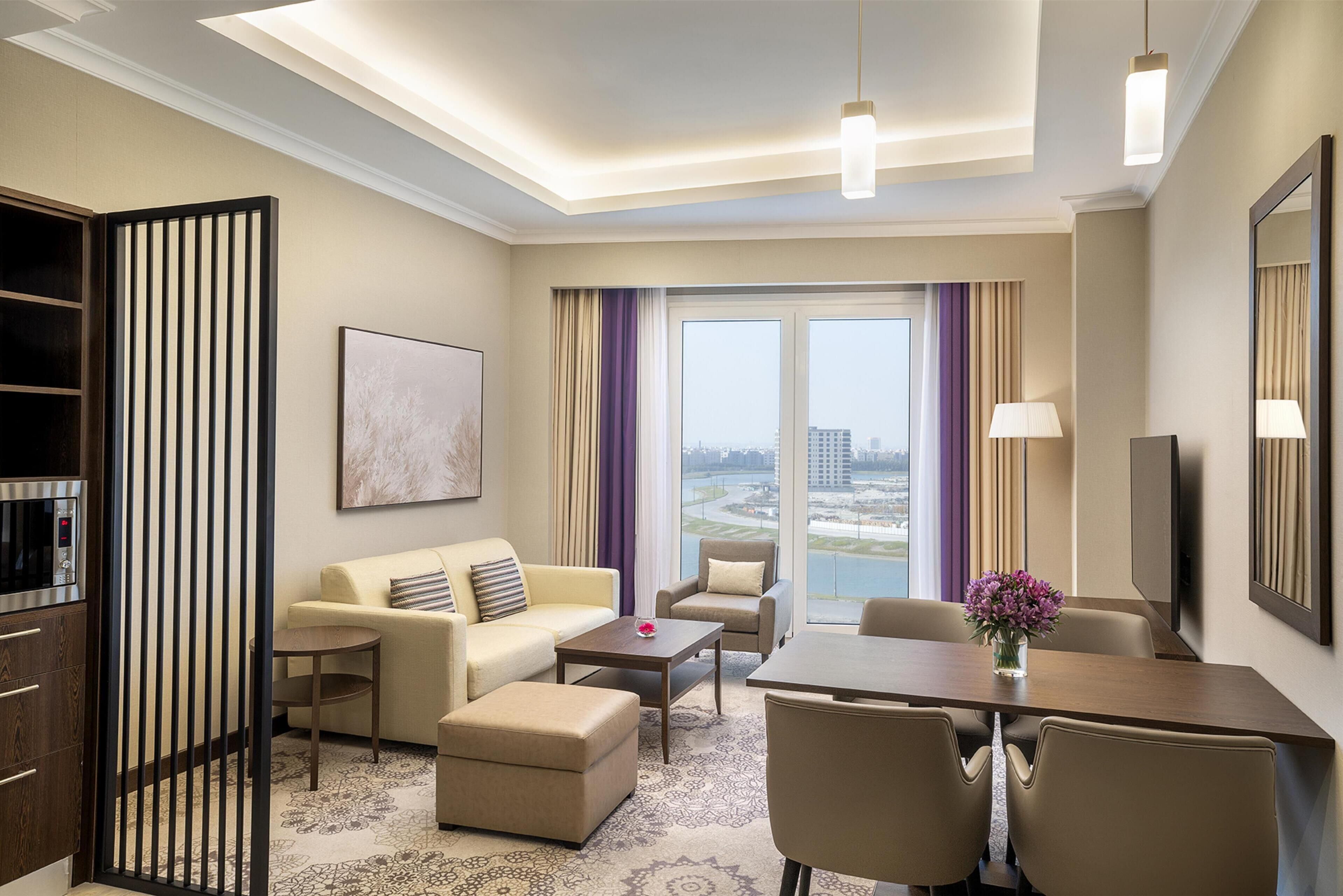 Une chambre d’hôtel moderne, dotée d’un canapé, de fauteuils, d’une table à manger et offrant une vue sur la ville grâce à de grandes fenêtres.