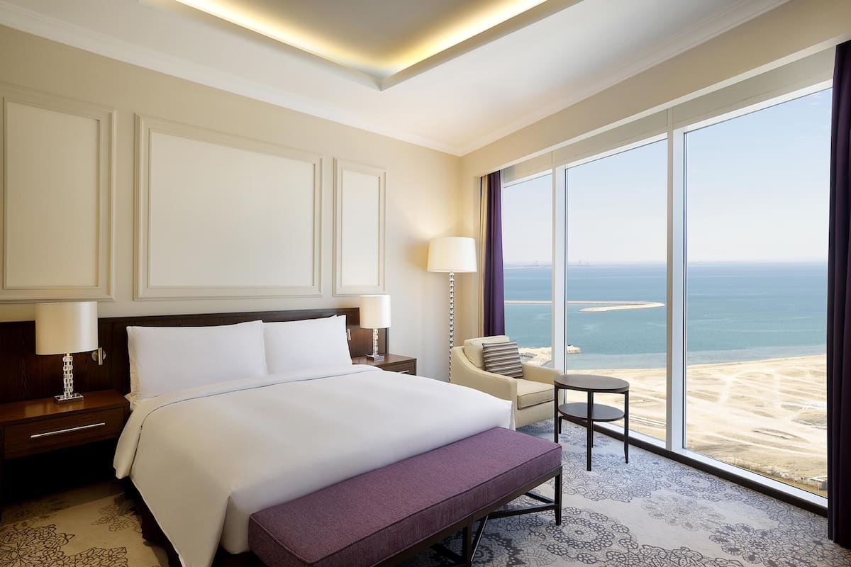 Quarto de hotel com uma cama grande, uma cadeira, uma mesinha e vista para a praia.