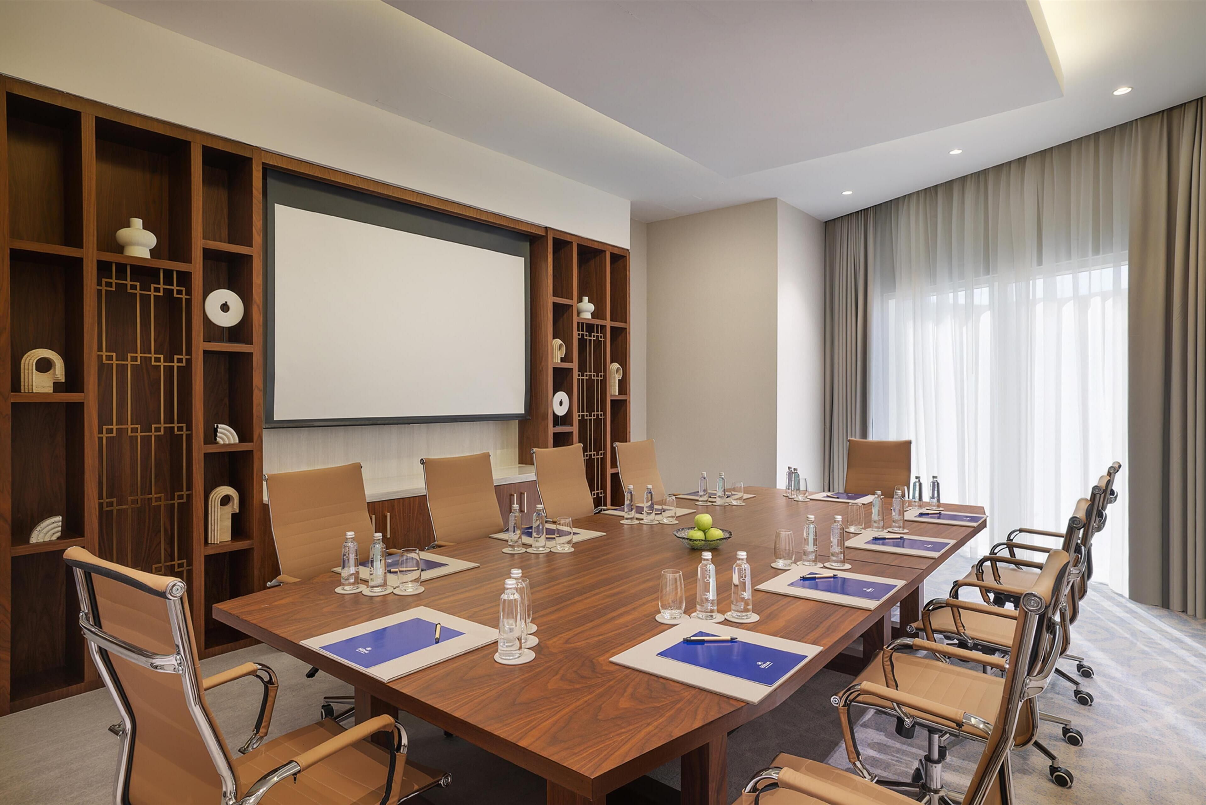 A conference room with a large wooden table, beige chairs, a projector screen, and a large window with curtains.